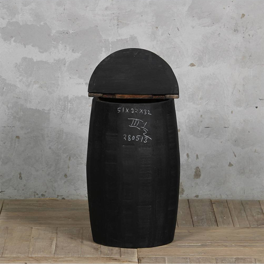 A black molded plastic or rubber trash can with a domed lid, standing on a light wood plank floor against a textured gray concrete wall. White handwritten markings are visible on the side of the container.