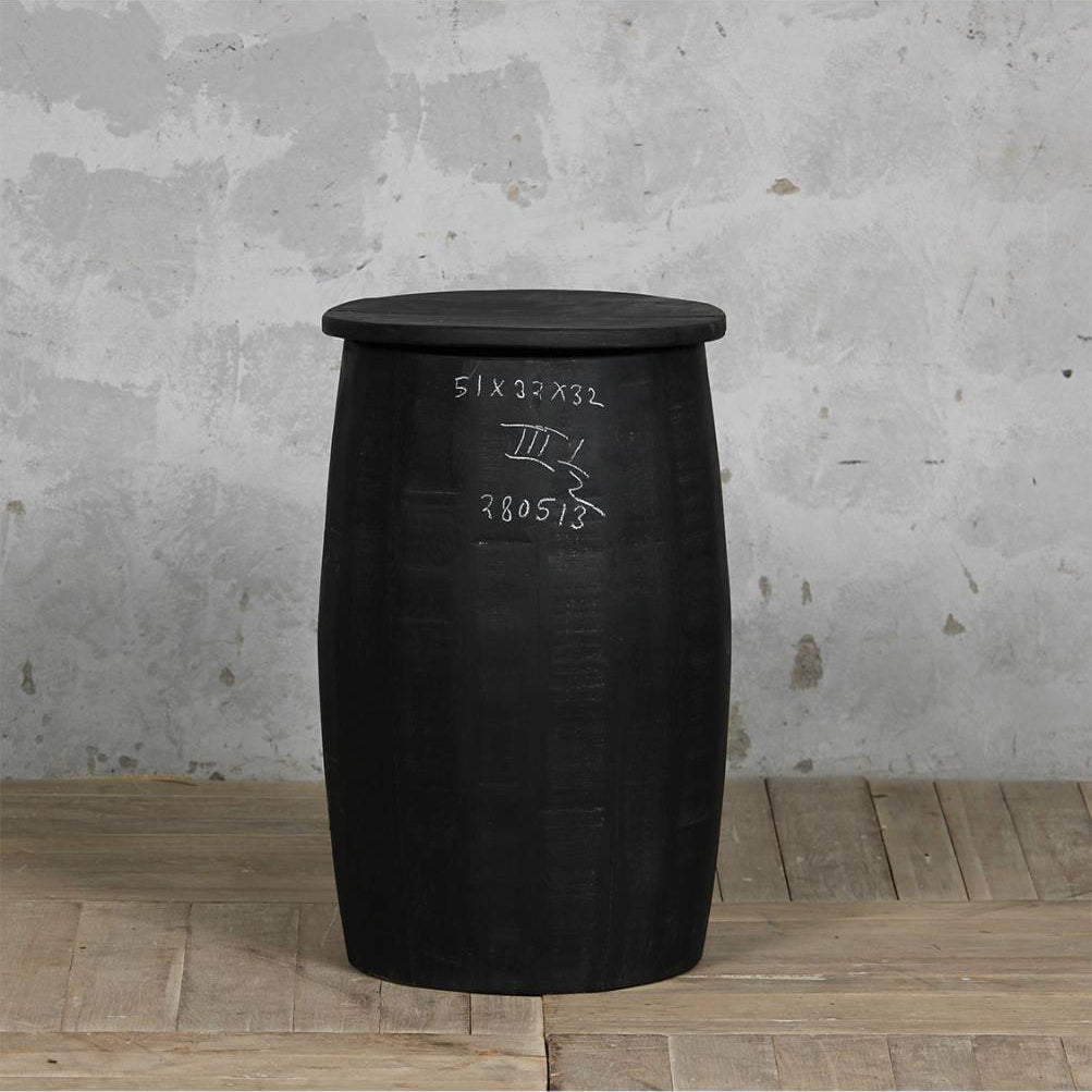A black barrel-shaped side table or stool with a flat circular top, standing on a light wood floor against a textured gray concrete wall. White handwritten markings are visible on the front.
