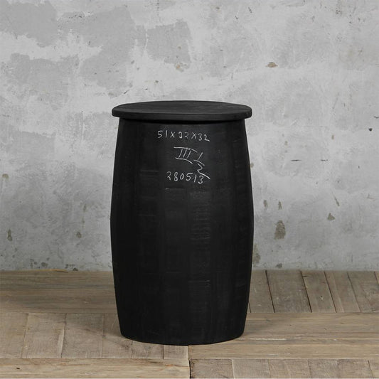 A black barrel-shaped side table or stool with a flat circular top, standing on a light wood floor against a textured gray concrete wall. White handwritten markings are visible on the front.