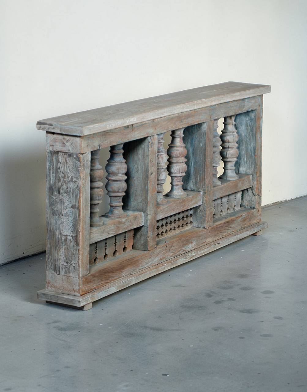 A rustic, weathered wooden console table or bench with carved balustrade details and a distressed gray-blue and brown finish, set against a plain off-white wall and gray concrete floor.