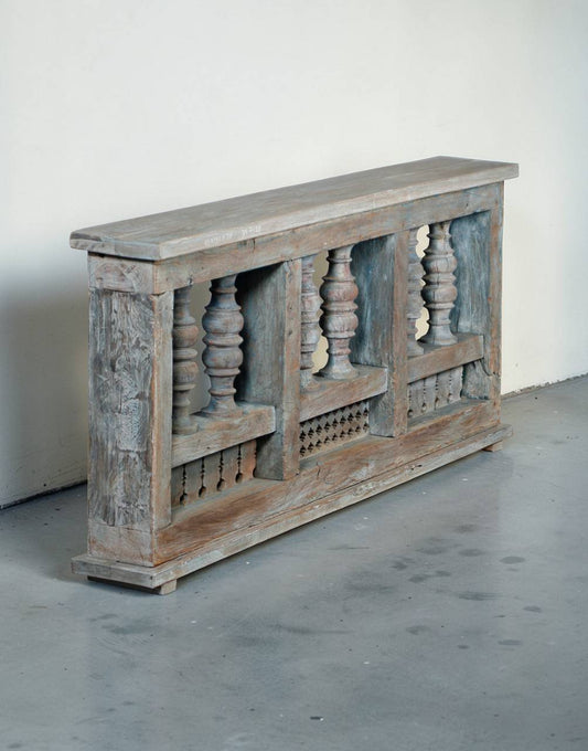 A rustic, weathered wooden console table or bench with carved balustrade details and a distressed gray-blue and brown finish, set against a plain off-white wall and gray concrete floor.