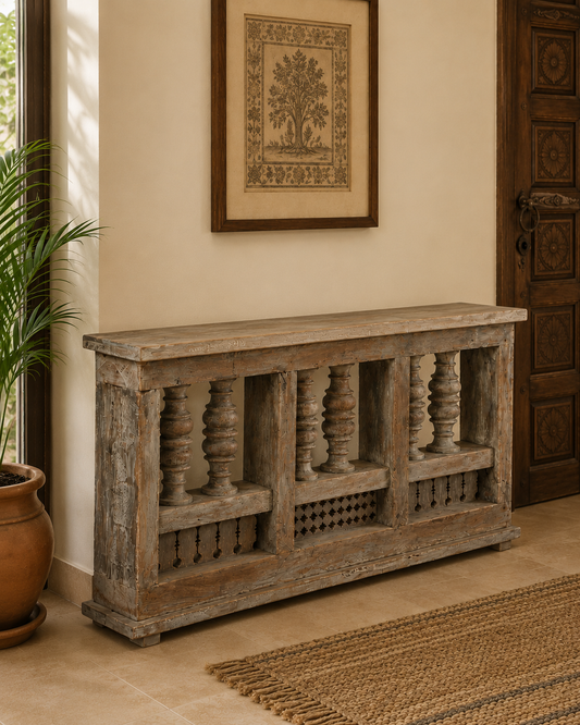 A rustic distressed wood entryway console table with carved balustrade-style legs, placed in a warm-toned room beside a potted plant, framed wall art, and a carved wooden door.