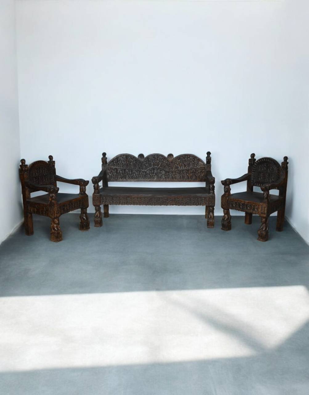A carved dark brown wooden sofa and two matching armchairs placed against a plain white wall in a minimalist room with a gray floor.