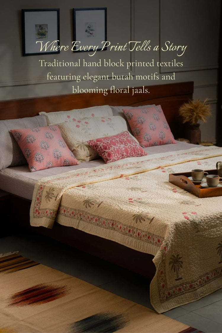 A styled bedroom scene showing a bed made up with a cream block-printed quilt or bedspread, pink patterned pillows, and matching decorative pillows. 