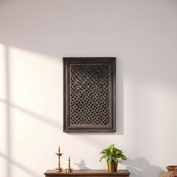 A rectangular dark carved wooden screen panel with an intricate lattice pattern, leaning against a white wall on a concrete floor.
