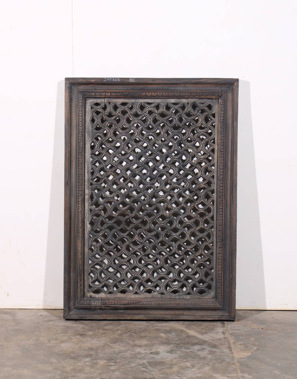 A rectangular dark carved wooden screen panel with an intricate lattice pattern, leaning against a white wall on a concrete floor.