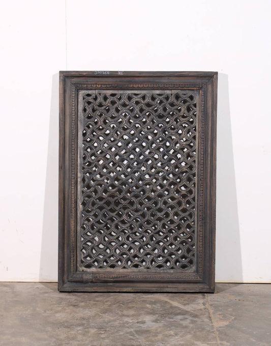 A rectangular dark carved wooden screen panel with an intricate lattice pattern, leaning against a white wall on a concrete floor.