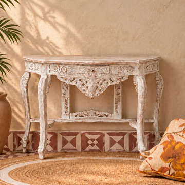 An ornate white carved console table with distressed detailing, set against a textured beige wall with leaf shadows, a patterned tile base, a woven rug, and a floral throw pillow.