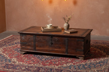 A dark brown wooden storage chest with metal hardware and weathered detailing, photographed in a warehouse setting with a gray concrete floor and white wall.