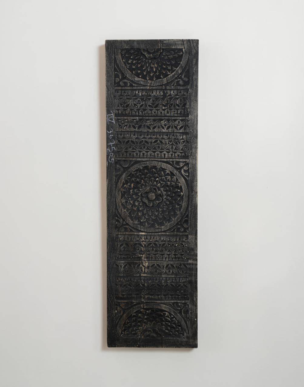 A tall, narrow black carved wooden plaque with intricate geometric and floral patterns, mounted on a plain white wall.