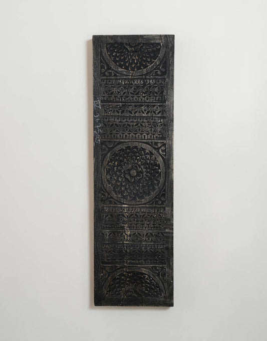A tall, narrow black carved wooden plaque with intricate geometric and floral patterns, mounted on a plain white wall.