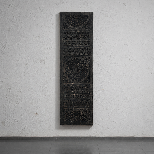 A tall, narrow dark carved wooden panel with intricate geometric and floral patterns, mounted on a textured white wall above a gray floor.