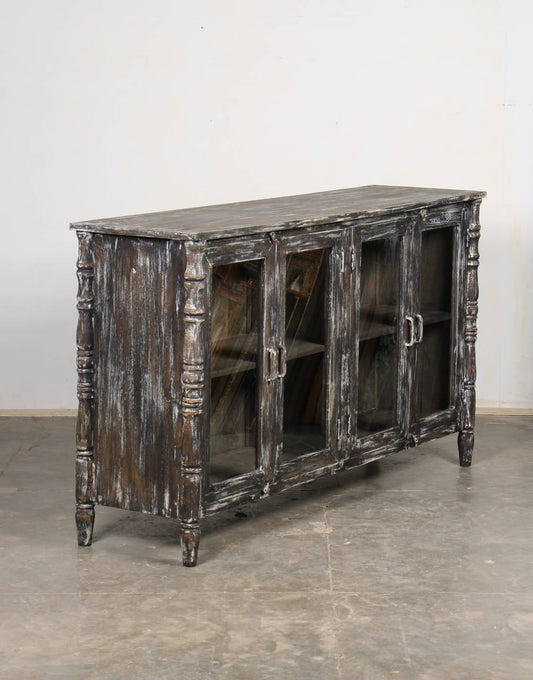 A distressed dark wood sideboard cabinet with glass doors, shown in a warehouse or studio setting with a concrete floor and plain wall.