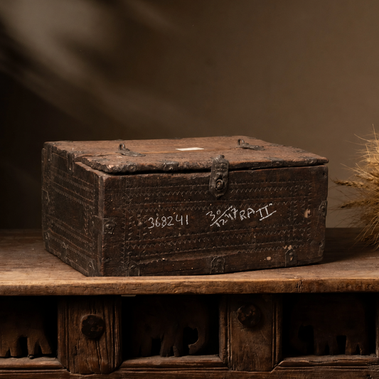 An antique dark brown wooden storage chest with metal latch hardware, resting on a matching rustic wooden cabinet against a dark brown background with soft leaf shadows.