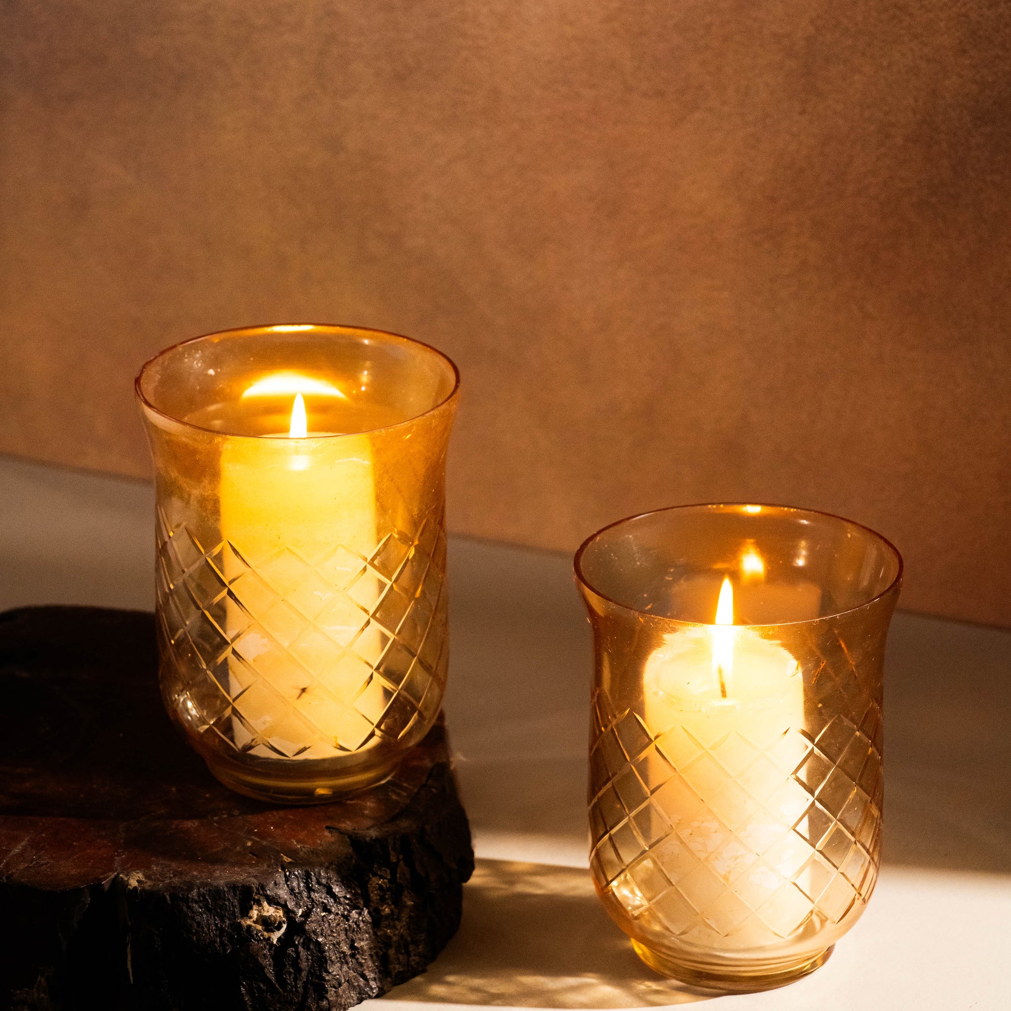 Two lit candles in textured glass holders on a wooden surface with a warm brown background.