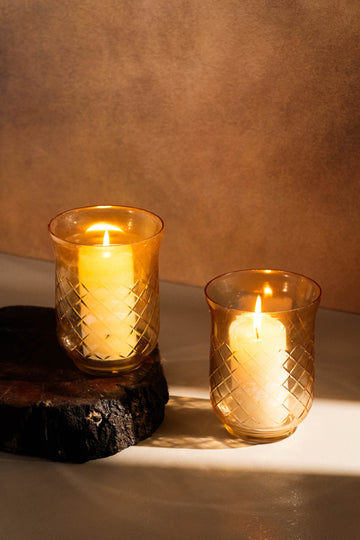 Two lit candles in textured glass holders on a wooden surface with a warm brown background.
