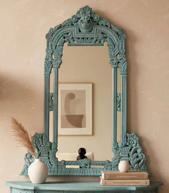 An ornate turquoise painted wooden console table with a matching arched mirror, styled in a warm beige room with a floor lamp and wood herringbone flooring.