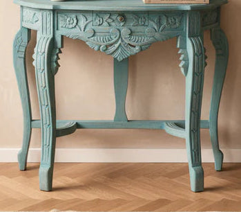 An ornate turquoise painted wooden console table with a matching arched mirror, styled in a warm beige room with a floor lamp and wood herringbone flooring.