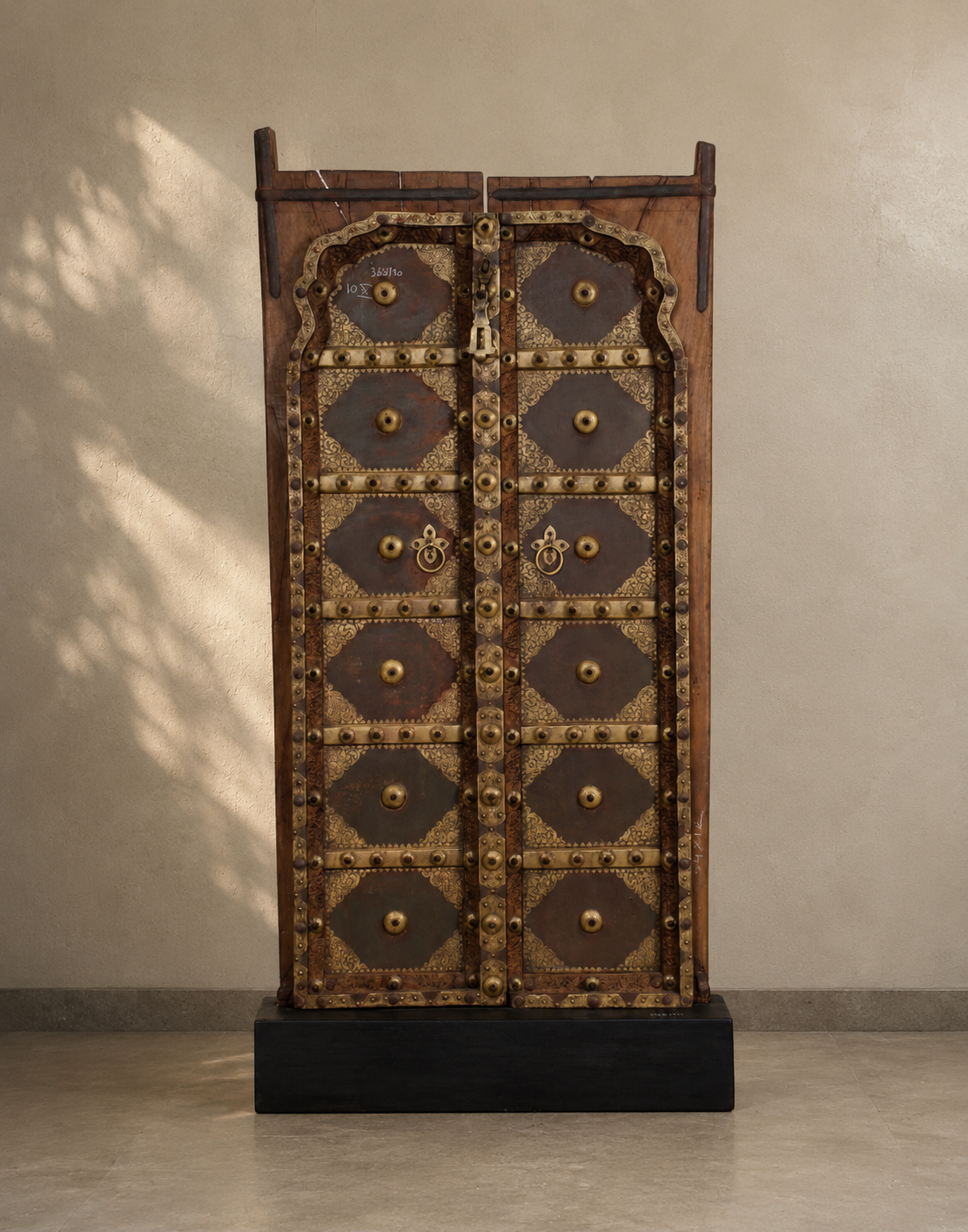 A tall, narrow traditional wooden cabinet with ornate metal detailing and multiple diamond-shaped door panels, displayed on a black base against a textured beige wall.