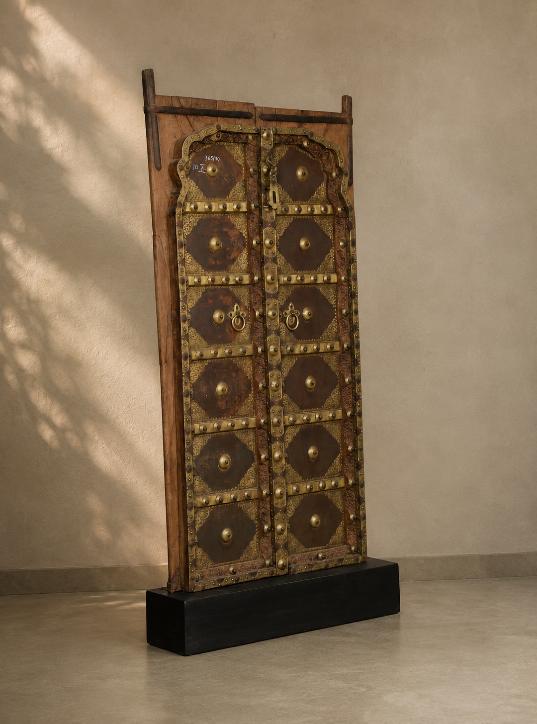 A tall, narrow traditional wooden door with ornate brass hardware and decorative panels, displayed on a black pedestal against a textured beige wall with soft shadow light filtering from the side.