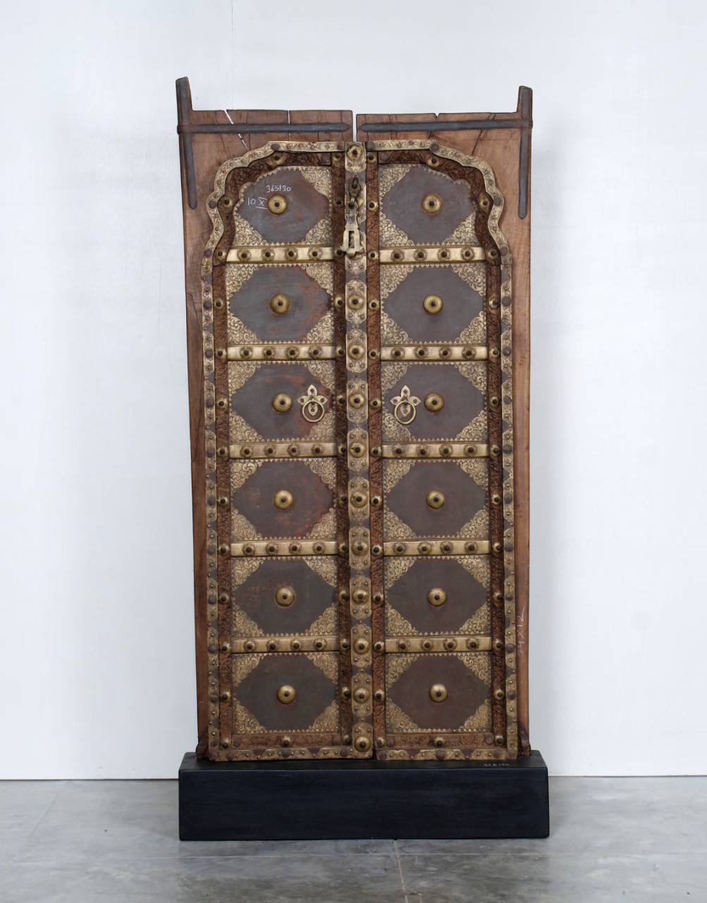 A tall, narrow wooden cabinet or door with ornate metal studs, decorative panels, and a lock, standing on a black base against a plain white wall.