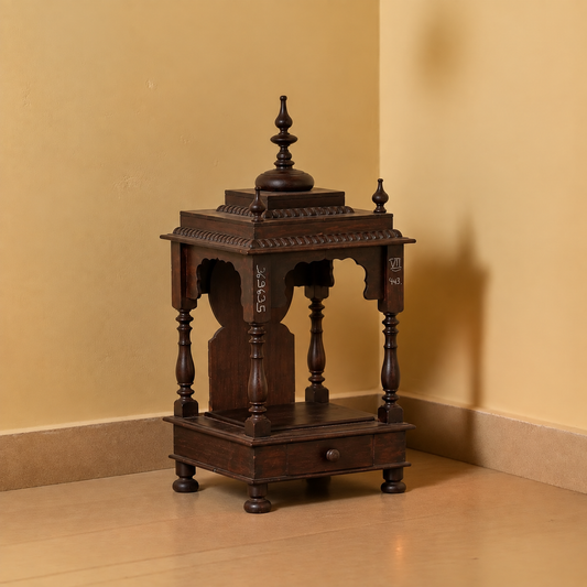 A dark brown carved wooden miniature altar or display stand with a single drawer and decorative finials, photographed in a corner with beige walls and floor.
