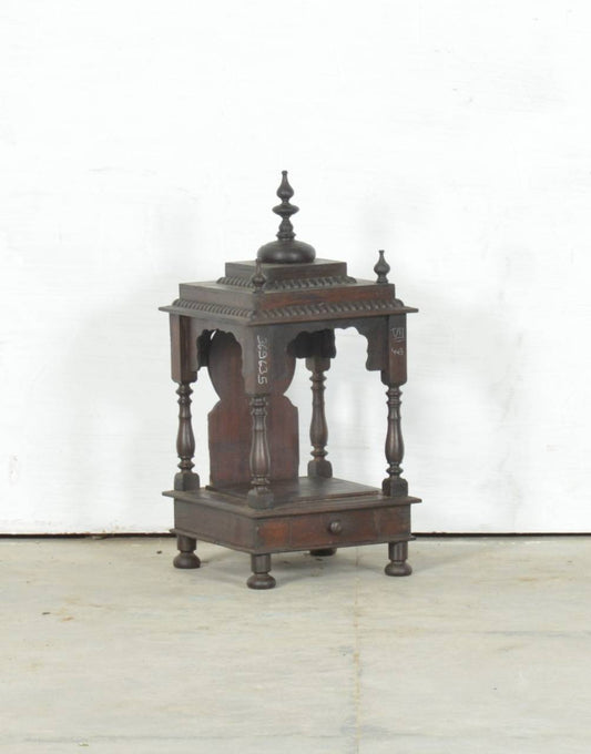 An antique dark brown wooden Shiva mandir or small temple structure with carved details, pedestal base, and a finial on top, photographed against a plain white wall and light floor.