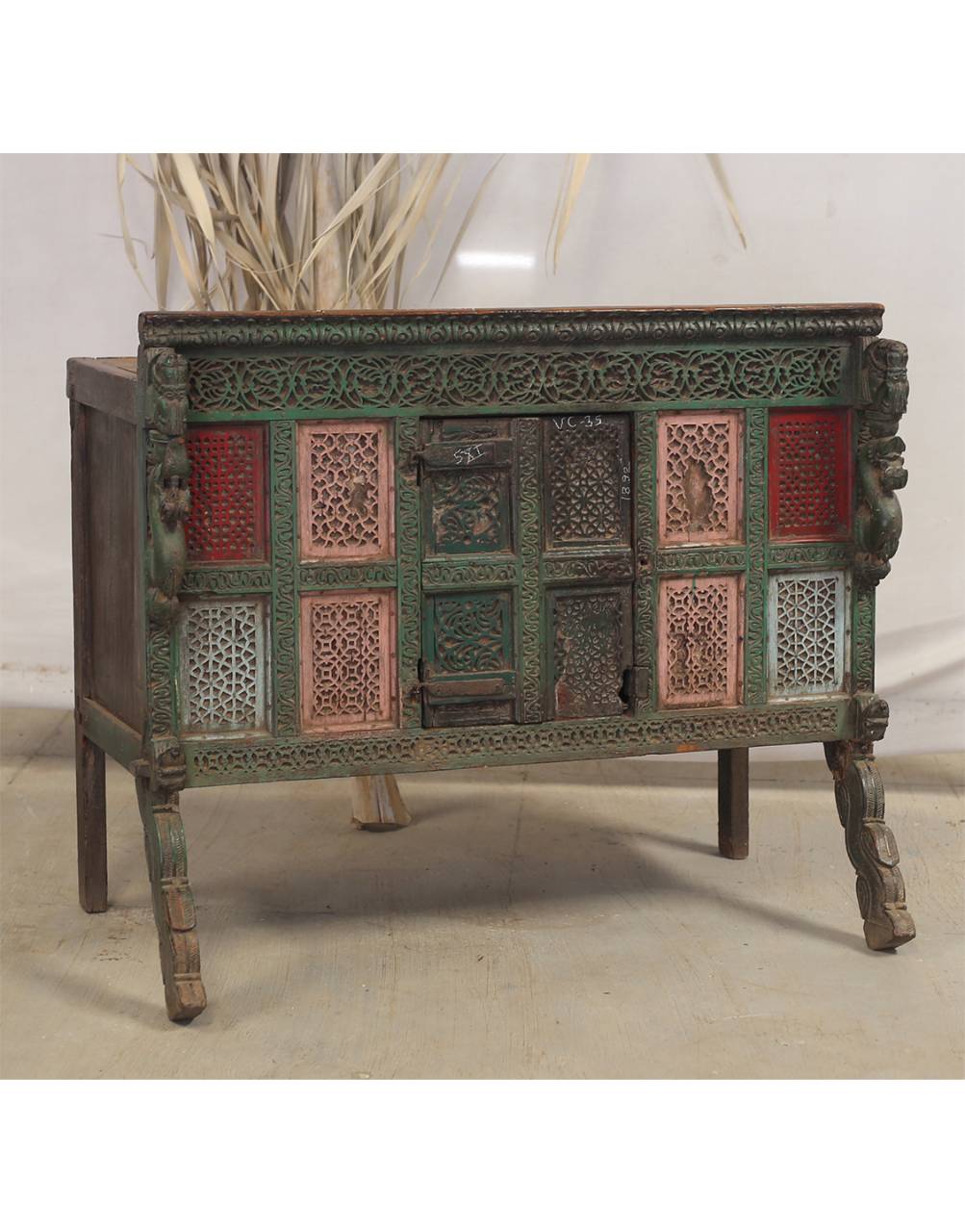 An ornate antique-style wooden cabinet or side table with carved details, a distressed green and brown finish, and decorative red and pink panels on the front, shown against a neutral studio backdrop.