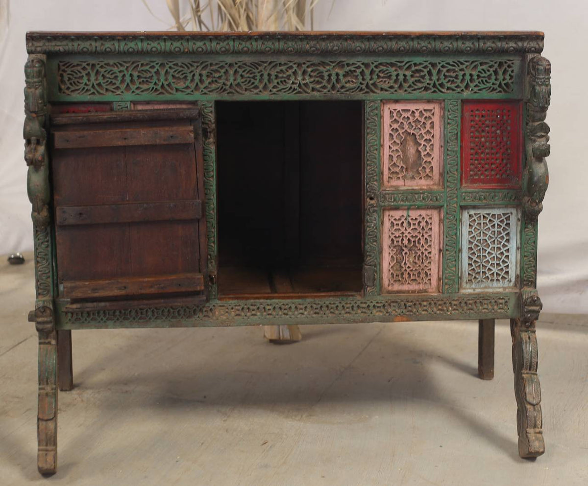 An antique carved wooden cabinet or side table with a distressed green-painted finish, featuring an open central compartment, a dark wooden door on the left, and decorative pink, red, and blue carved panels on the right.