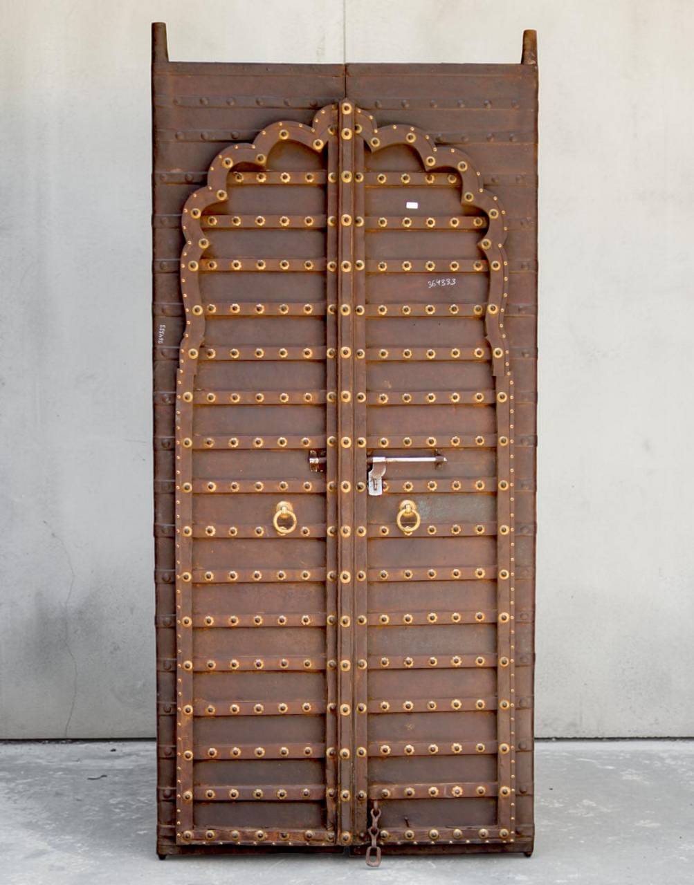 A tall, narrow wooden door panel with an arched top and extensive brass-colored metal studs and decorative accents, photographed upright against a plain light gray wall and concrete floor.