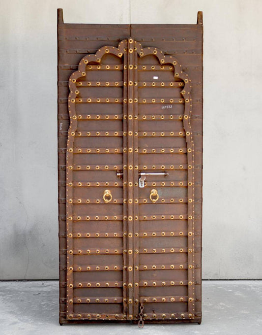 A tall, narrow wooden door panel with an arched top and extensive brass-colored metal studs and decorative accents, photographed upright against a plain light gray wall and concrete floor.