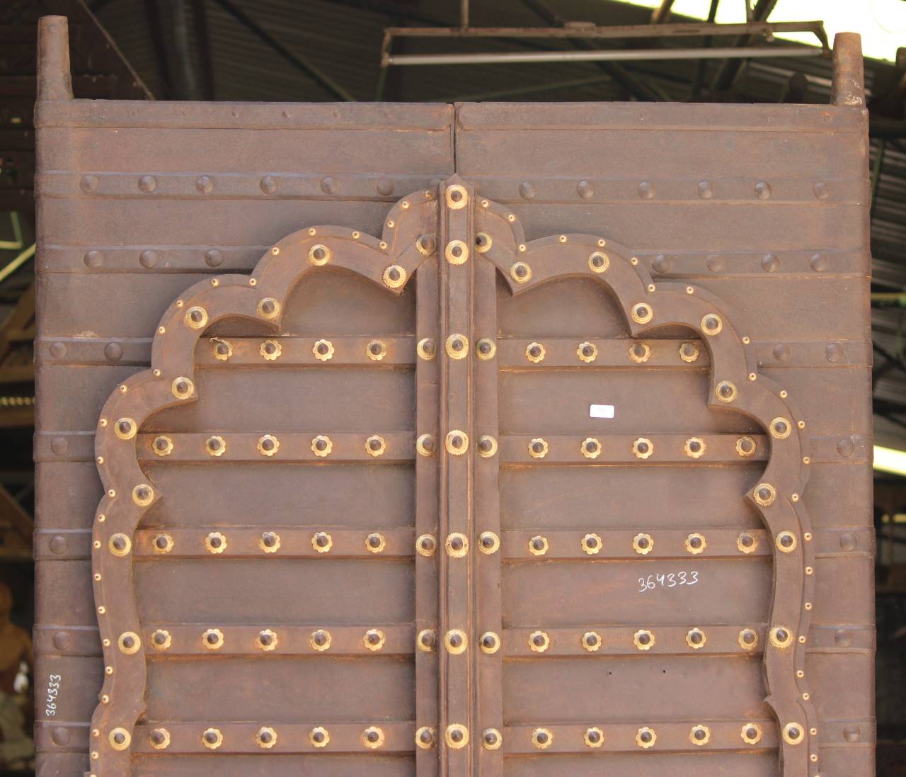 A pair of large, rustic brown steel doors with ornate scalloped edges and rows of decorative brass studs, photographed in an industrial warehouse setting.