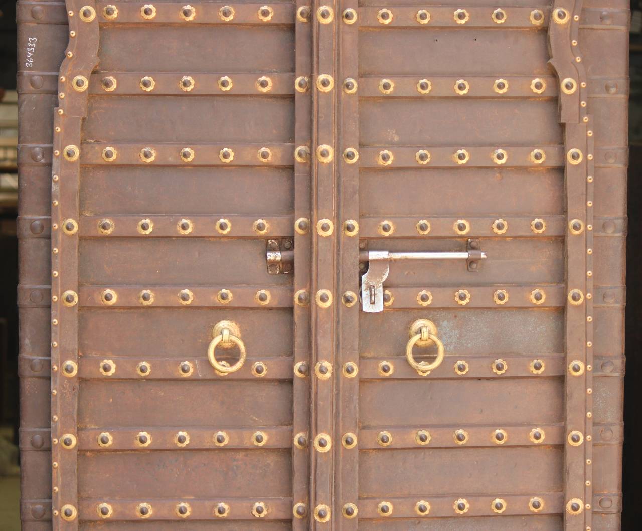 Close-up of a rustic brown metal door with rows of round brass studs, a central vertical panel, a silver latch, and two hanging ring handles.