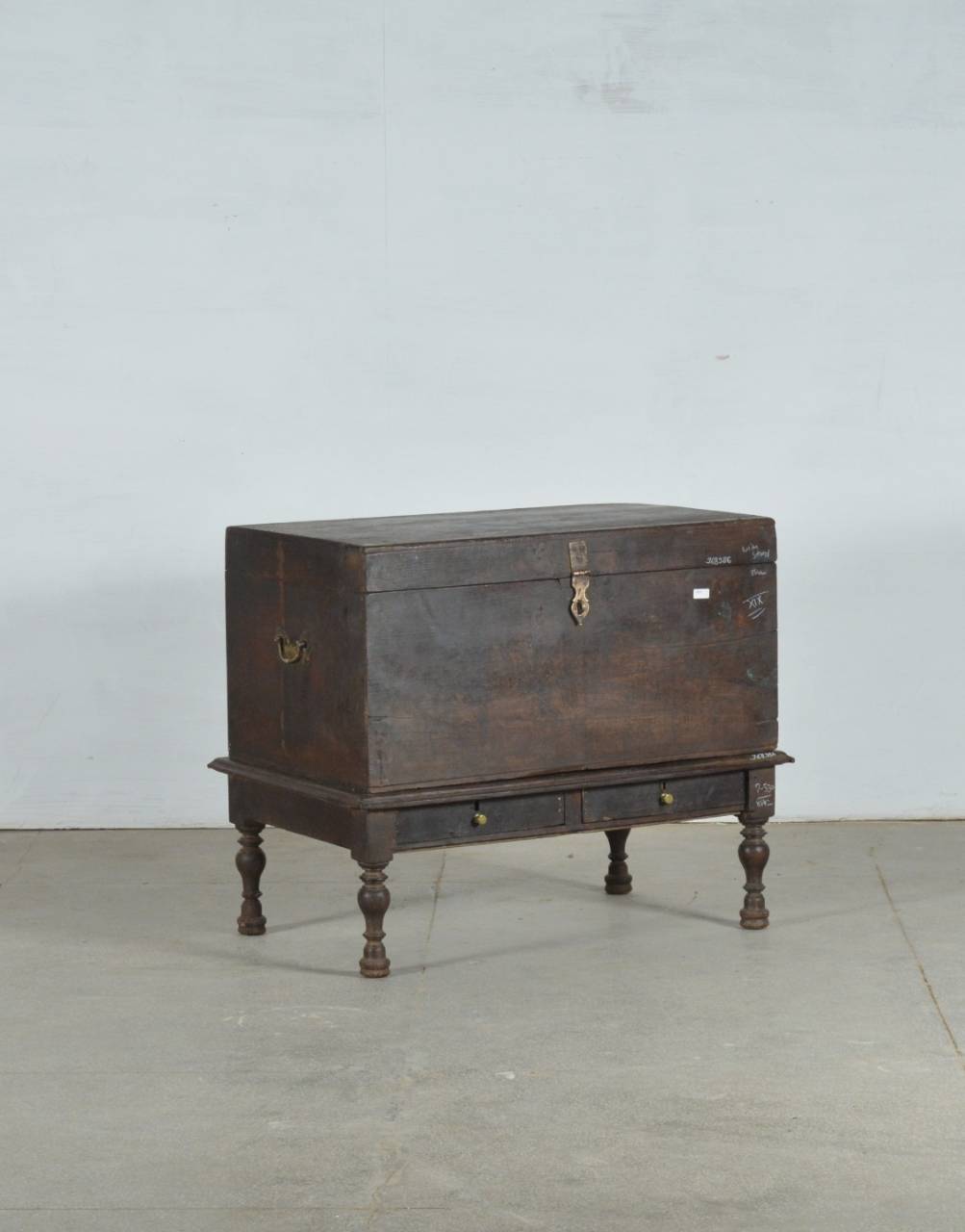 An antique dark brown wooden chest with a raised base, turned legs, and brass hardware, photographed against a plain light gray wall and concrete floor.