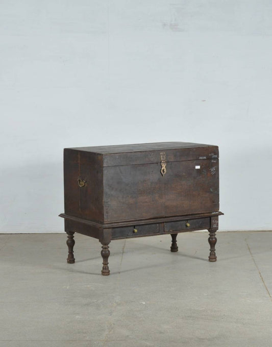 An antique dark brown wooden chest with a raised base, turned legs, and brass hardware, photographed against a plain light gray wall and concrete floor.