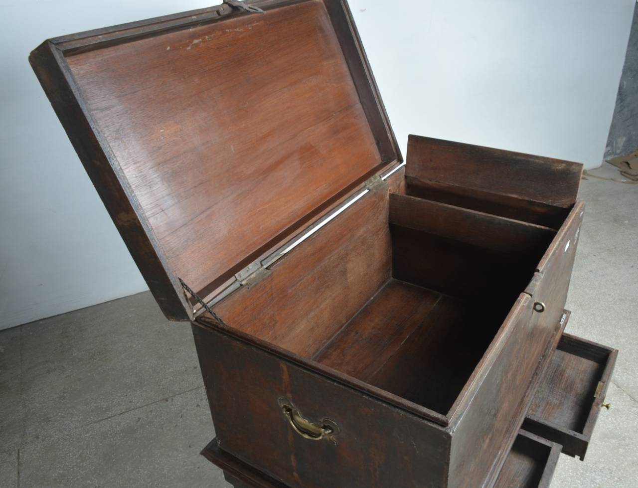 A dark brown wooden chest with an open lid, showing an interior organizer with compartments and a drawer, photographed in a warehouse setting.