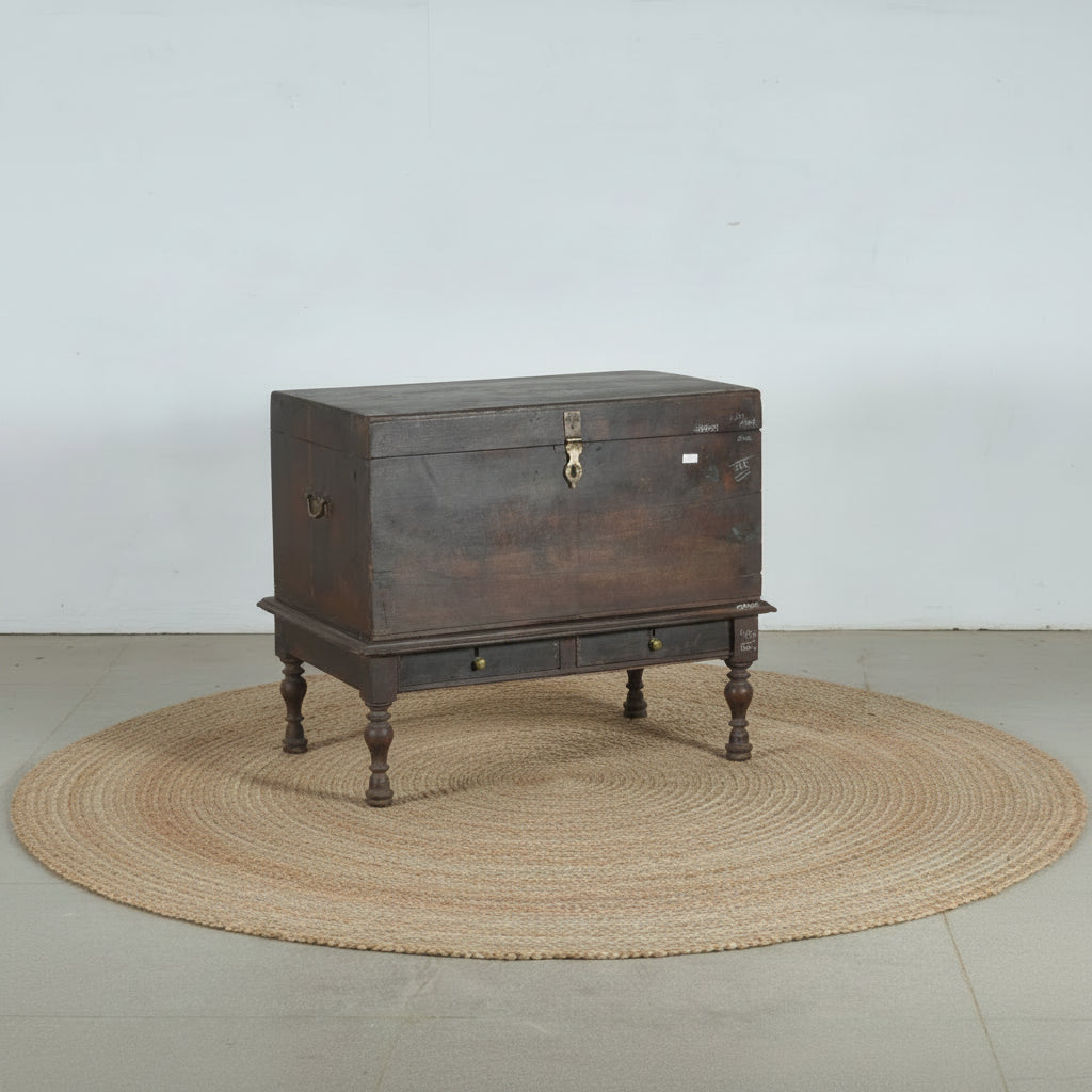 An antique dark brown wooden chest with a raised base, turned legs, and brass hardware, photographed against a plain light gray wall and concrete floor.