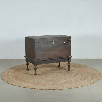 An antique dark brown wooden chest with a raised base, turned legs, and brass hardware, photographed against a plain light gray wall and concrete floor.