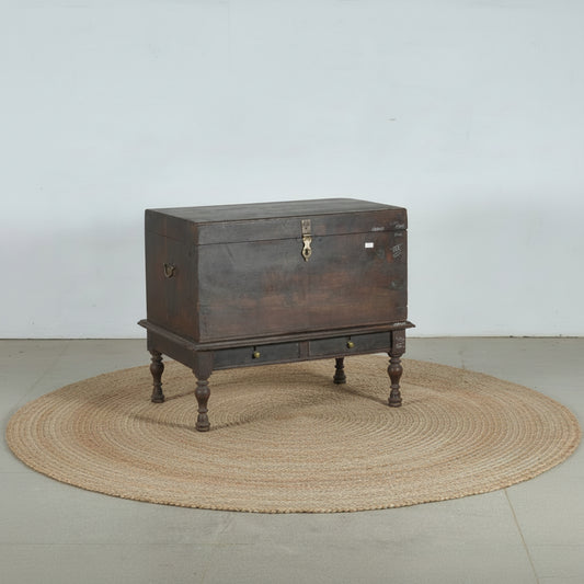 An antique dark brown wooden chest with a raised base, turned legs, and brass hardware, photographed against a plain light gray wall and concrete floor.
