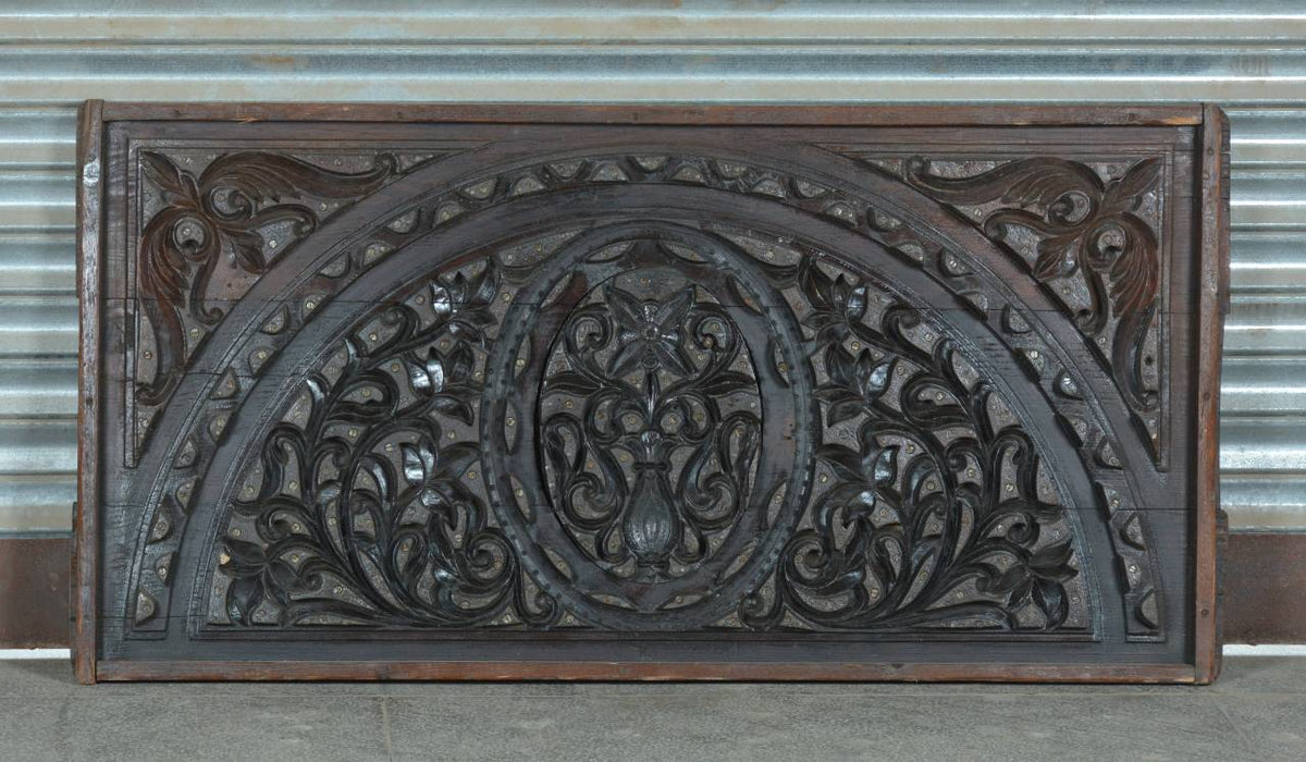 An ornate carved wooden panel with a semi-circular arch design and intricate scrollwork, photographed against a corrugated metal wall.
