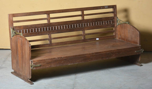 A vintage wooden daybed with a slatted backrest and footrest, featuring brass hardware, photographed in a warehouse setting against a yellow wall.