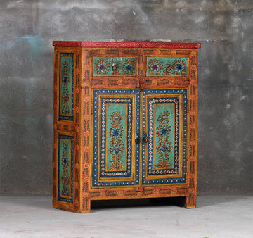 A hand-painted wooden cabinet with intricate floral patterns in turquoise, blue, and gold on a warm orange-brown base, set against a textured gray wall.