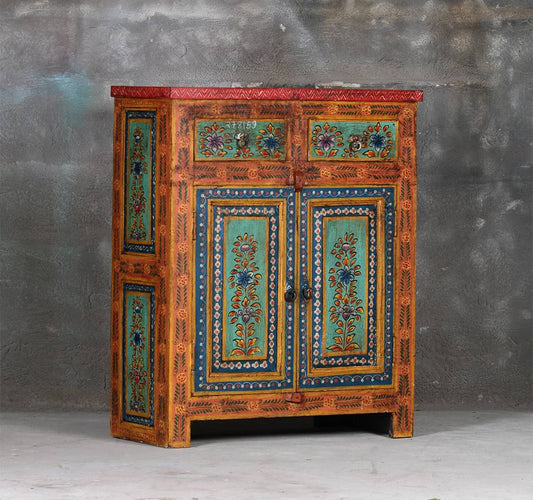 A hand-painted wooden cabinet with intricate floral patterns in turquoise, blue, and gold on a warm orange-brown base, set against a textured gray wall.