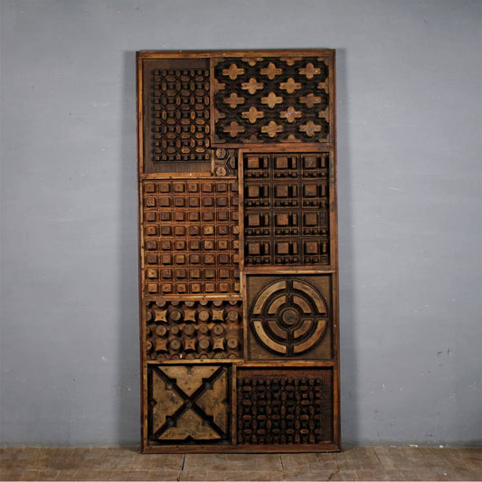A tall rectangular wooden decorative panel with multiple carved geometric and textured square panels, photographed against a plain gray wall and light floor.