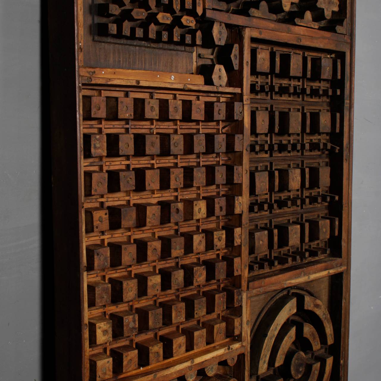An antique wooden compartment organizer cabinet with multiple small drawers, a lattice grid design, and a circular dial mechanism, photographed against a neutral gray background.