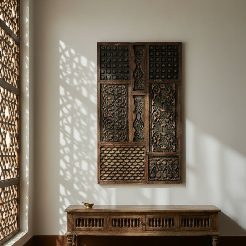 A large rectangular dark carved wooden wall panel with intricate geometric and floral patterns, mounted on a light beige wall next to a small potted plant.