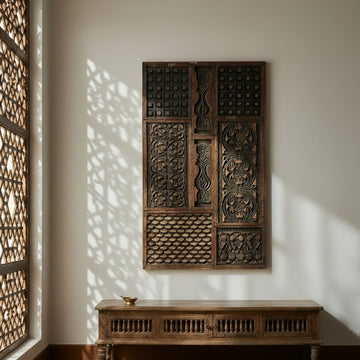 A large rectangular dark carved wooden wall panel with intricate geometric and floral patterns, mounted on a light beige wall next to a small potted plant.