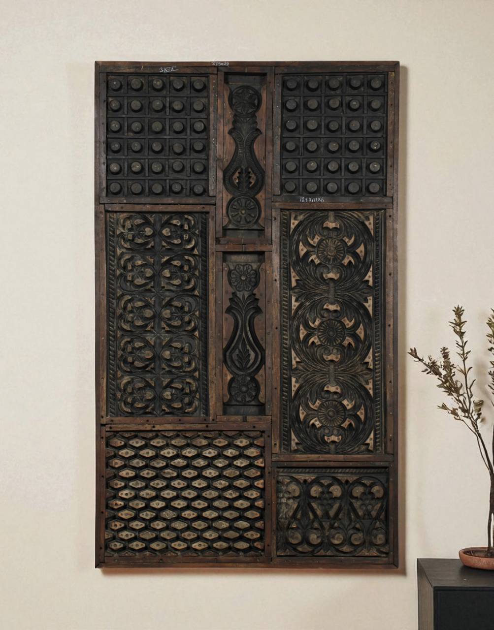 A large rectangular dark carved wooden wall panel with intricate geometric and floral patterns, mounted on a light beige wall next to a small potted plant.