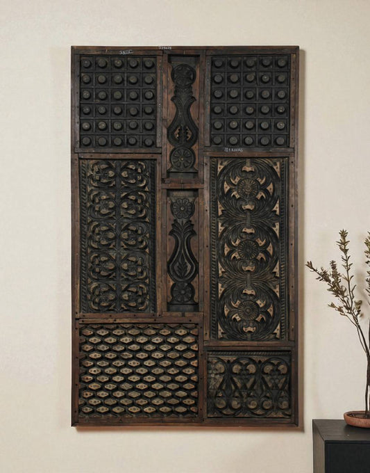 A large rectangular dark carved wooden wall panel with intricate geometric and floral patterns, mounted on a light beige wall next to a small potted plant.
