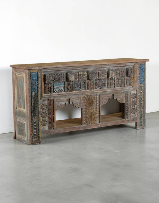 A rustic, hand-carved wooden console table with intricate geometric and floral patterns, featuring a weathered brown wood finish with blue and green painted accents, set against a plain white wall and gray concrete floor.